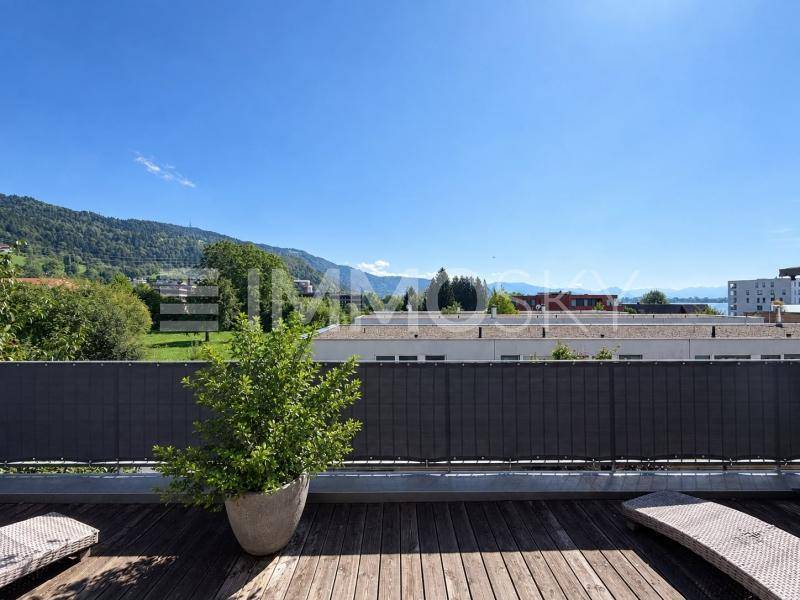 Terrasse mit Holzboden und weitem Blick auf Berge, See und grüne Landschaft unter strahlend blauem Himmel.