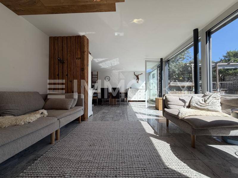 Helles Wohnzimmer mit großen Fensterfronten, modernen Sofas und einem Holzelement, das viel Tageslicht hereinlässt.