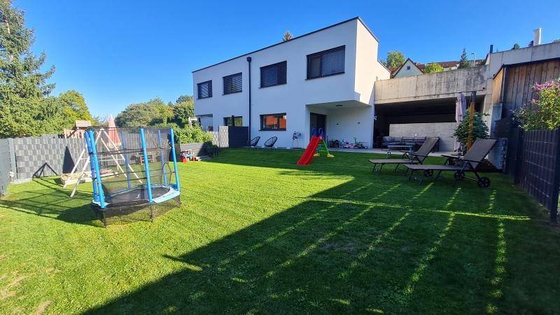 Moderne Hausfassade mit großem Garten, Rasenfläche und Kinderspielgeräten im Sonnenlicht.