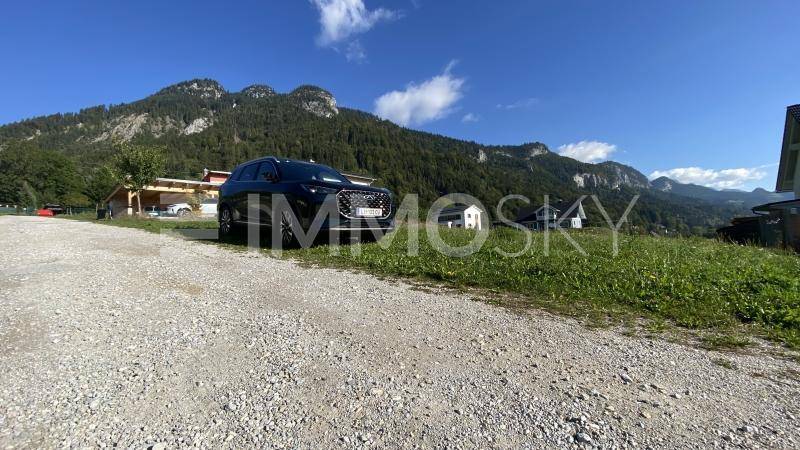 Schotterweg mit geparktem Auto vor einer beeindruckenden Bergkulisse und bewaldeten Hängen.