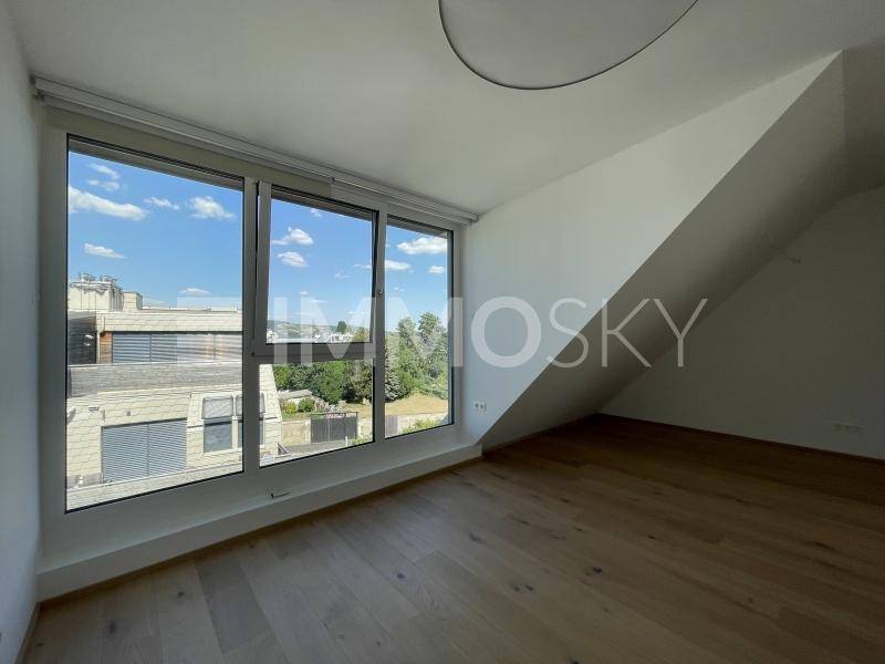 Helles Zimmer mit Holzboden, großen Fenstern und Blick auf die umliegende Bebauung und Natur.