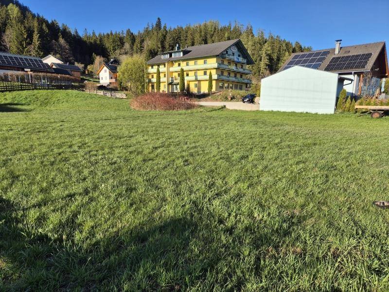 Sonnige Wiesenfläche mit Blick auf ein großes gelbes Gebäude und bewaldete Hügel.