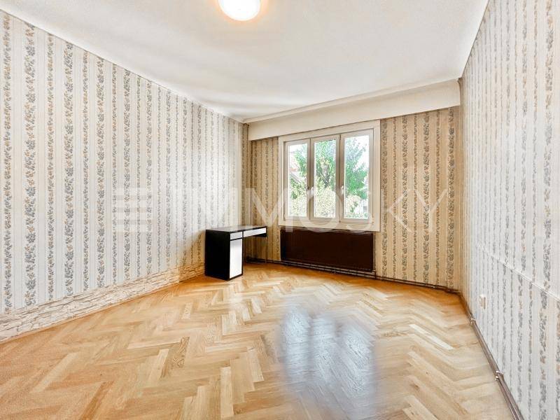 Geräumiges Zimmer mit gemusterter Tapete und Fischgrätparkett, bietet Platz für Schlaf- oder Arbeitsbereich.