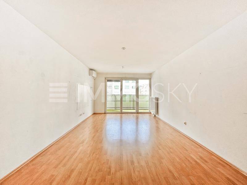 Leeres Wohnzimmer mit hellem Holzparkettboden, weißen Wänden und Zugang zum Balkon.
