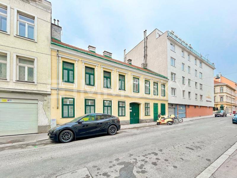 Straßenansicht eines historischen Gebäudes mit gelber Fassade und grünen Fensterläden in urbaner Umgebung.