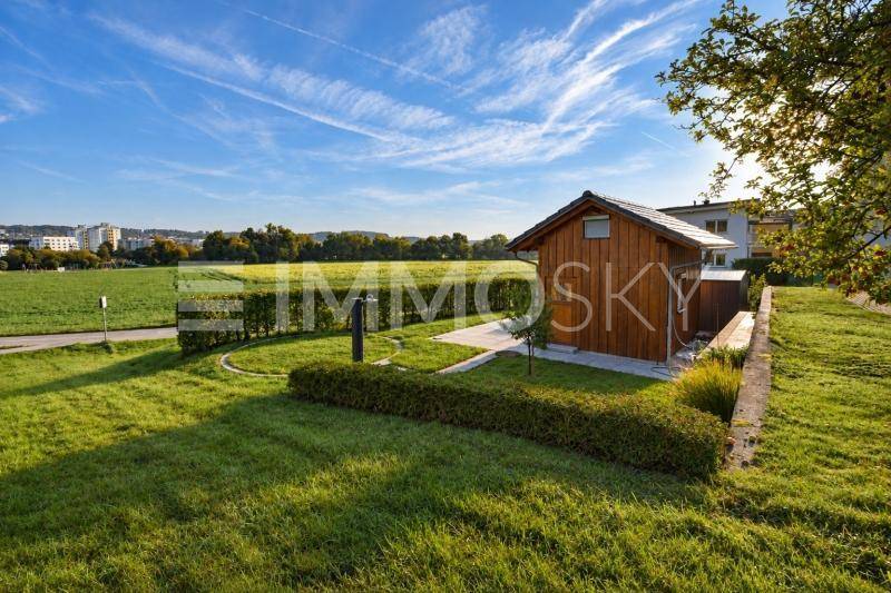 Sonnenbeschienener Außenbereich mit einem Holzschuppen, gepflegtem Rasen und weitem Blick in die Natur.