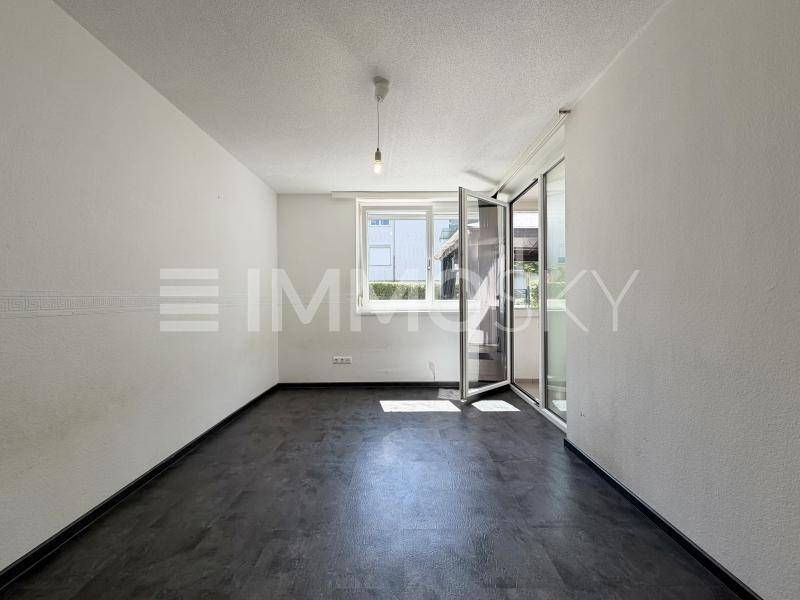 Helles, leeres Zimmer mit dunklem Bodenbelag, Fenster und Zugang zu einem Außenbereich.