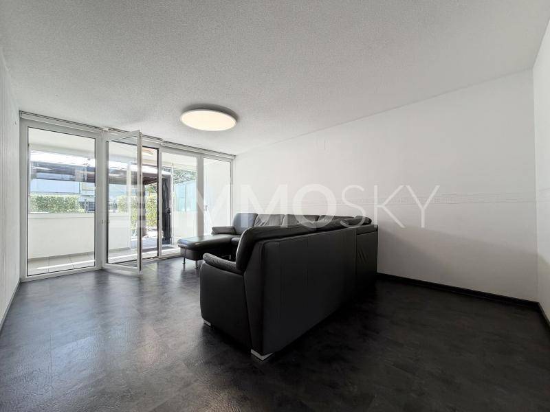 Geräumiges Wohnzimmer mit dunklem Sofa, modernem Bodenbelag und Zugang zu einem Balkon.