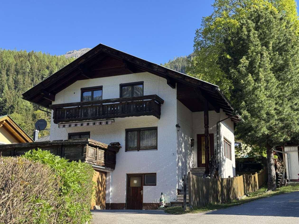 Freistehendes Haus mit Holzbalkonen und Satteldach, umgeben von Bäumen und Bergen unter blauem Himmel.