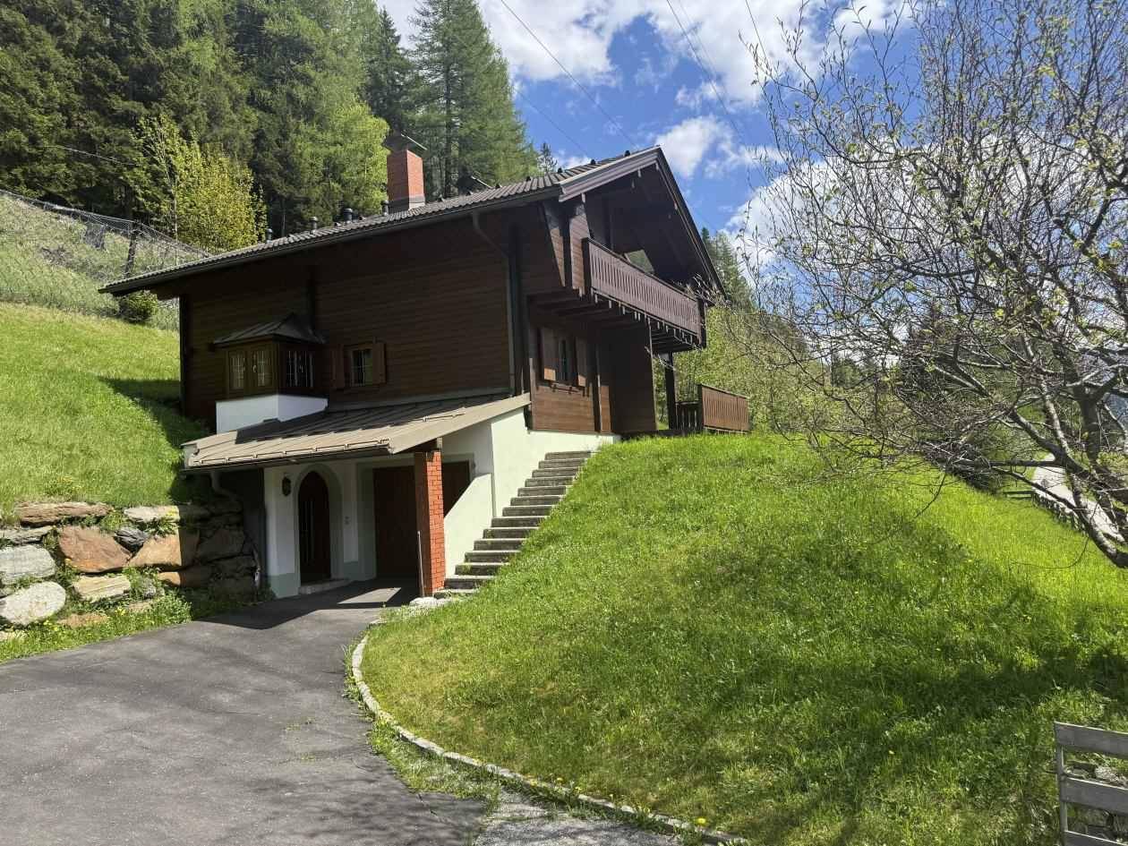 Traditionelles Holzhaus im Chalet-Stil am Hang mit Balkon und Carport, umgeben von grüner Natur.