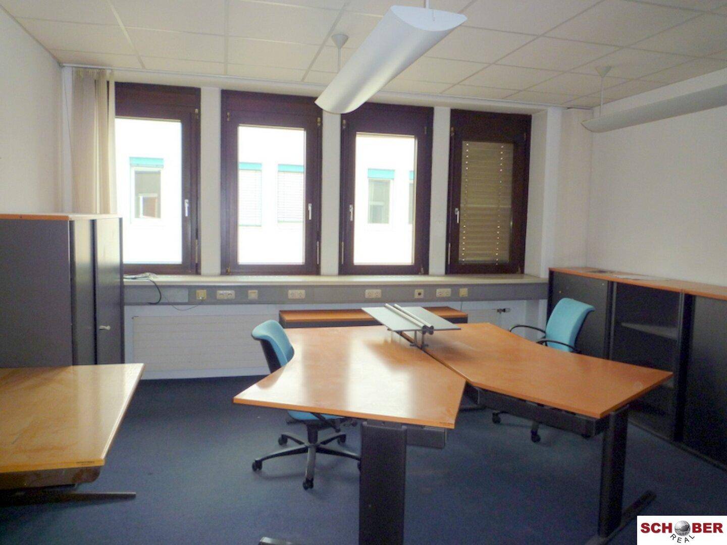Geräumiges Büro mit mehreren Arbeitsplätzen, Teppichboden und großen Fenstern.