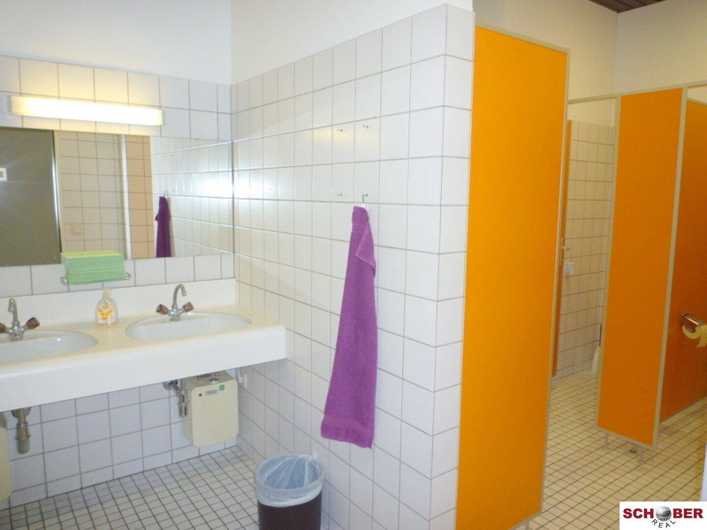 Sanitärbereich mit Doppelwaschbecken, Spiegel und orangefarbenen Trennwänden für Toilettenkabinen.