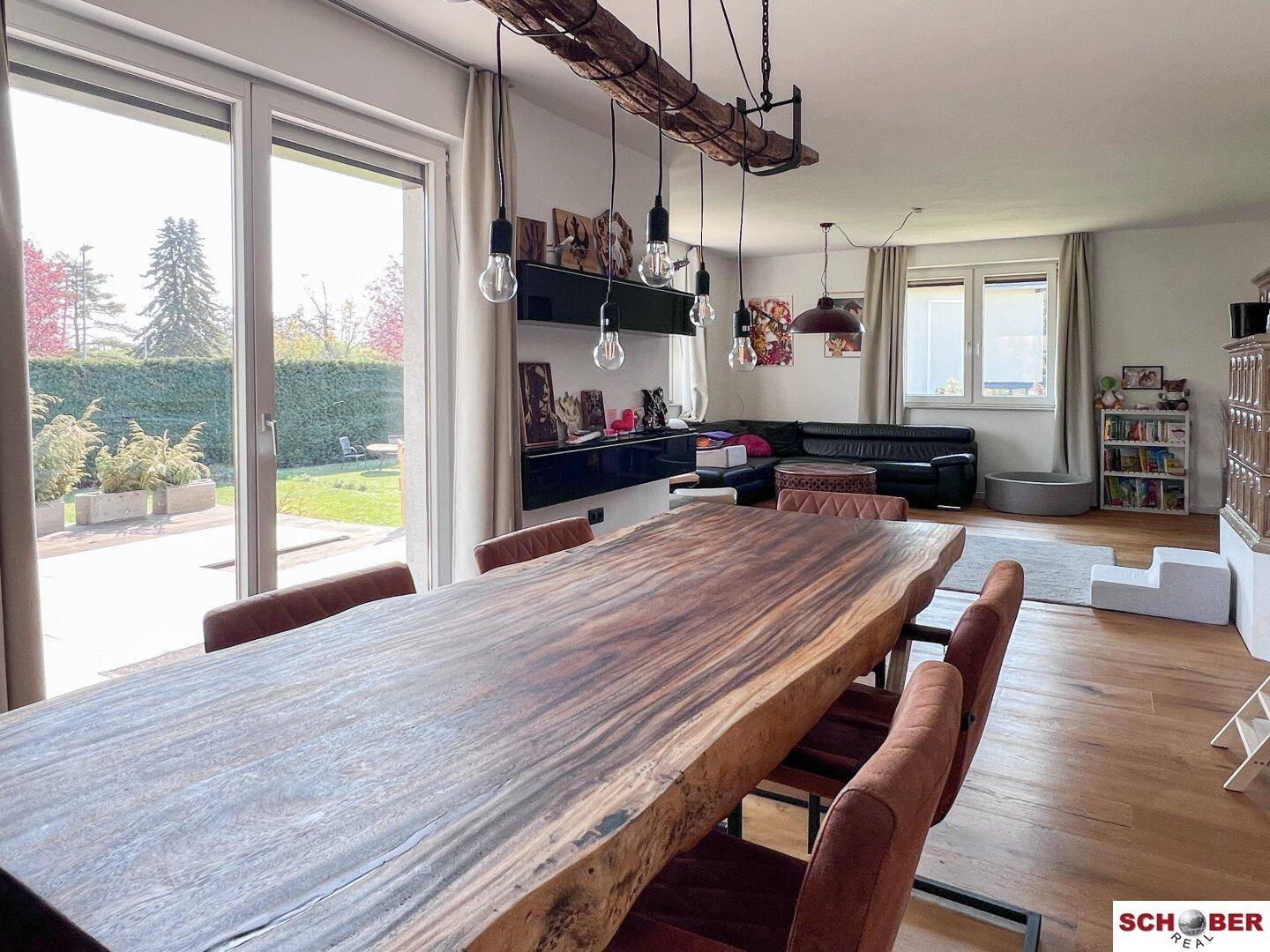 Lichtdurchfluteter Wohn-Essbereich mit großen Fenstern, Holztisch und Zugang zum Garten.