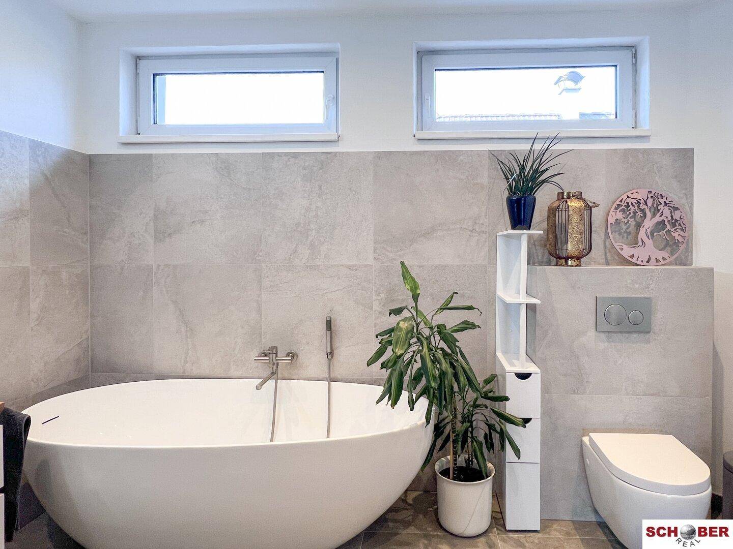 Helles Badezimmer mit freistehender Badewanne, wandhängendem WC und zwei Fenstern.