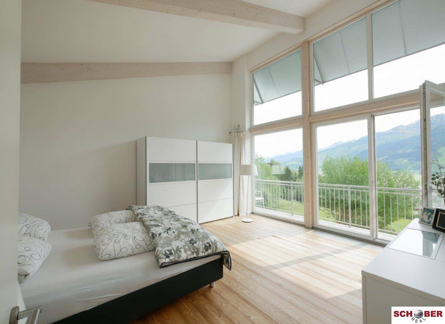 Schlafzimmer mit Holzboden, großen Fenstern und Balkonzugang mit Bergblick.
