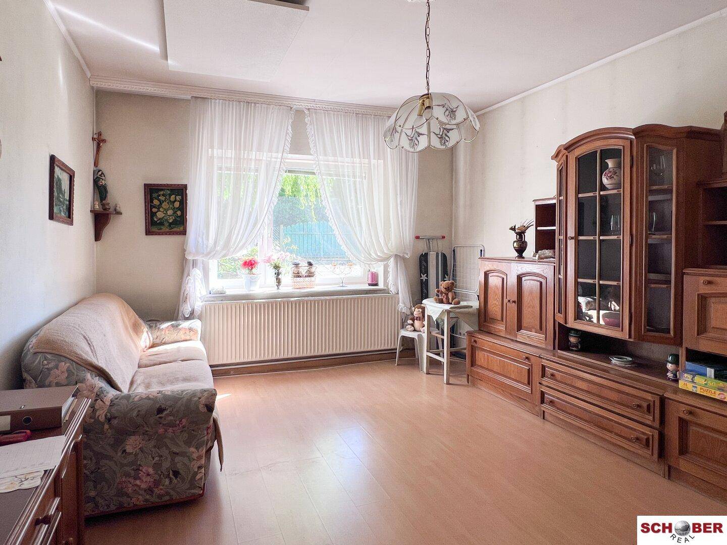 Helles Wohnzimmer mit Sofa, Holzschrank und großem Fenster mit Vorhängen.