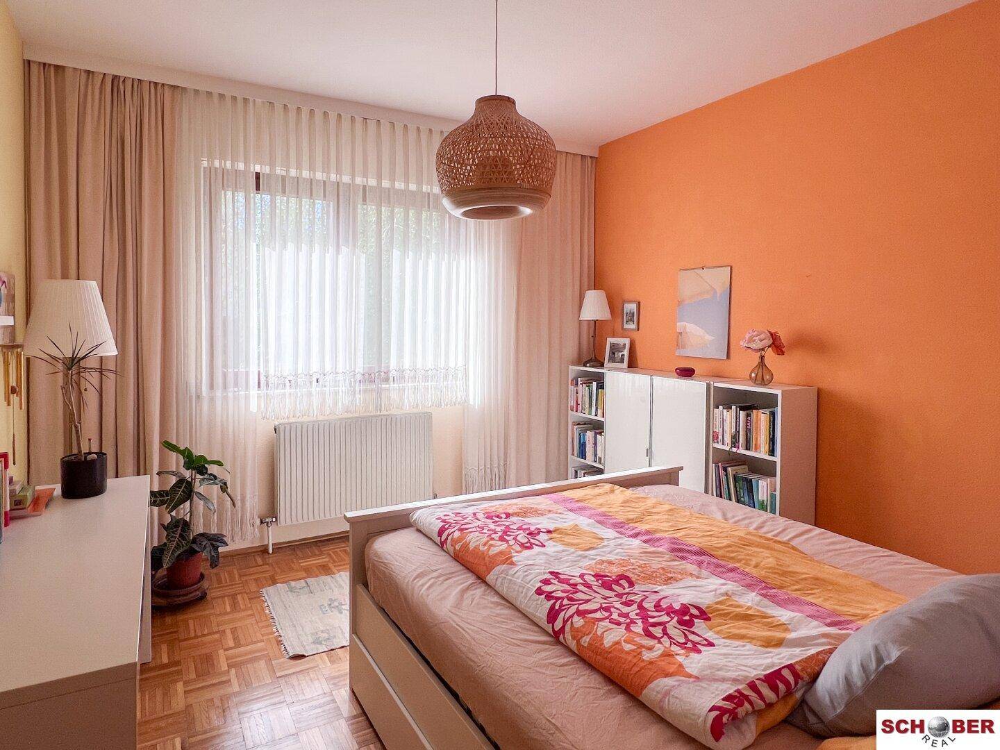 Schlafzimmer mit orangefarbener Akzentwand, Parkettboden und großem Fenster für viel Licht.