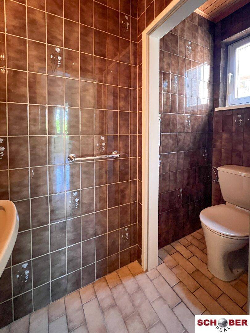 Kleines Gäste-WC mit braunen Fliesen, Toilette, Waschbecken und praktischem Handlauf am Duschbereich.