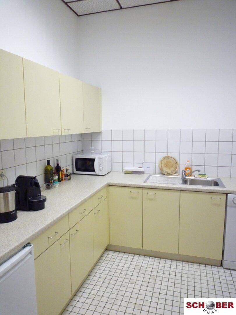 Kleine Teeküche mit hellen Schränken, Spüle, Mikrowelle und Fliesenboden.