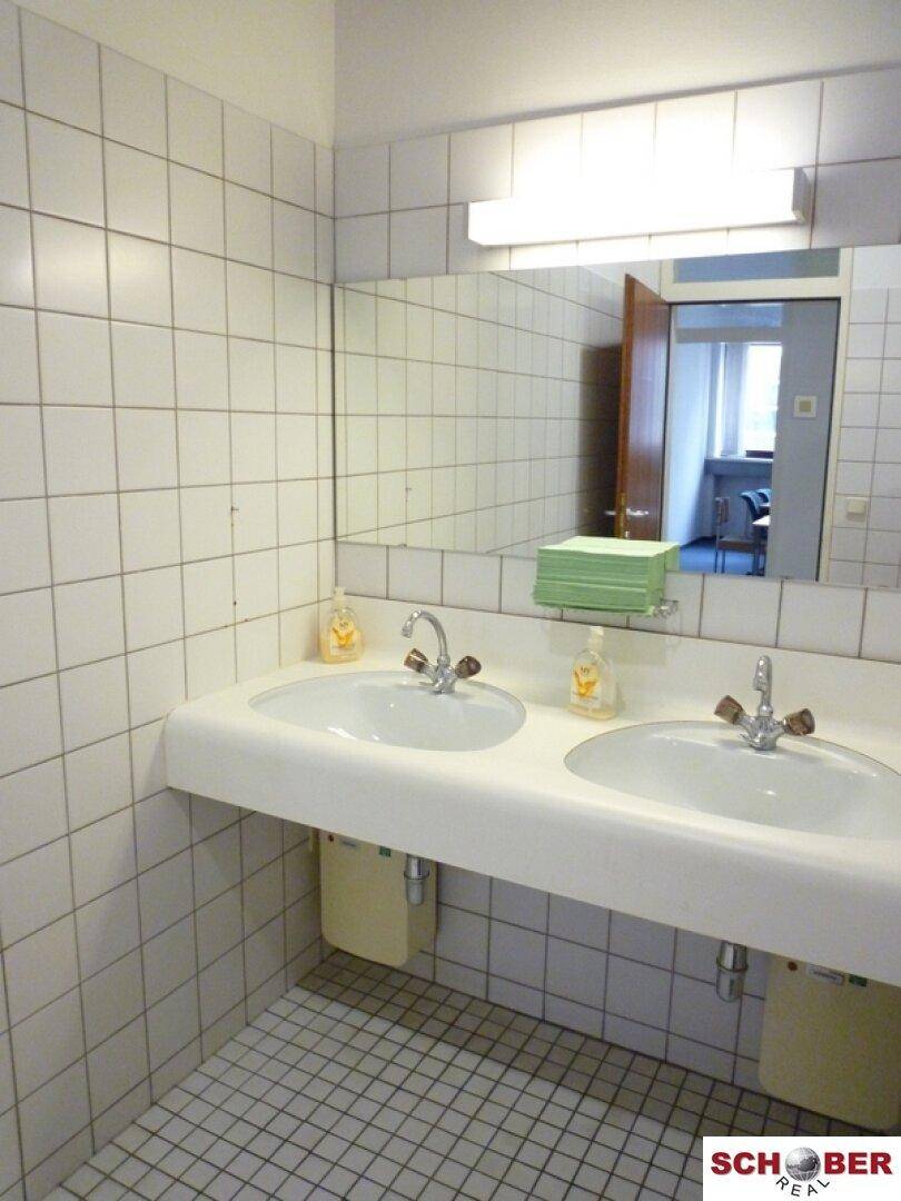 Waschraum mit zwei Waschbecken, großem Spiegel und weißen Fliesen an Wänden und Boden.