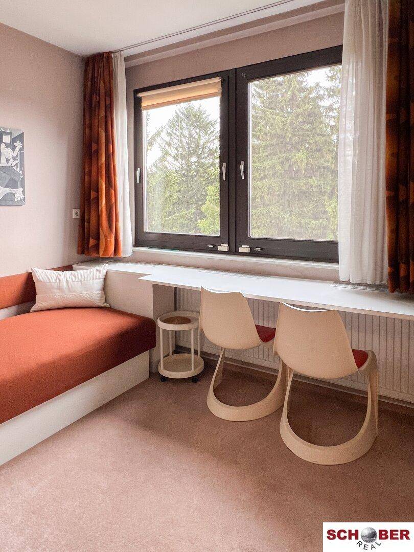 Schlaf- oder Arbeitszimmer mit Einzelbett, Schreibtisch und großem Fenster mit Blick ins Grüne.