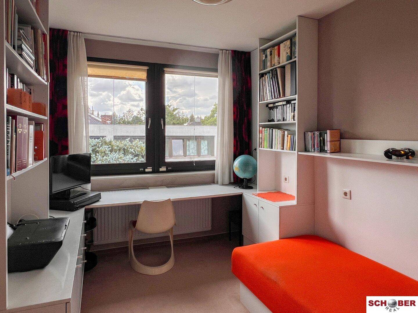Schlaf- oder Arbeitszimmer mit Einzelbett, Schreibtisch, Bücherregalen und einem Fenster.