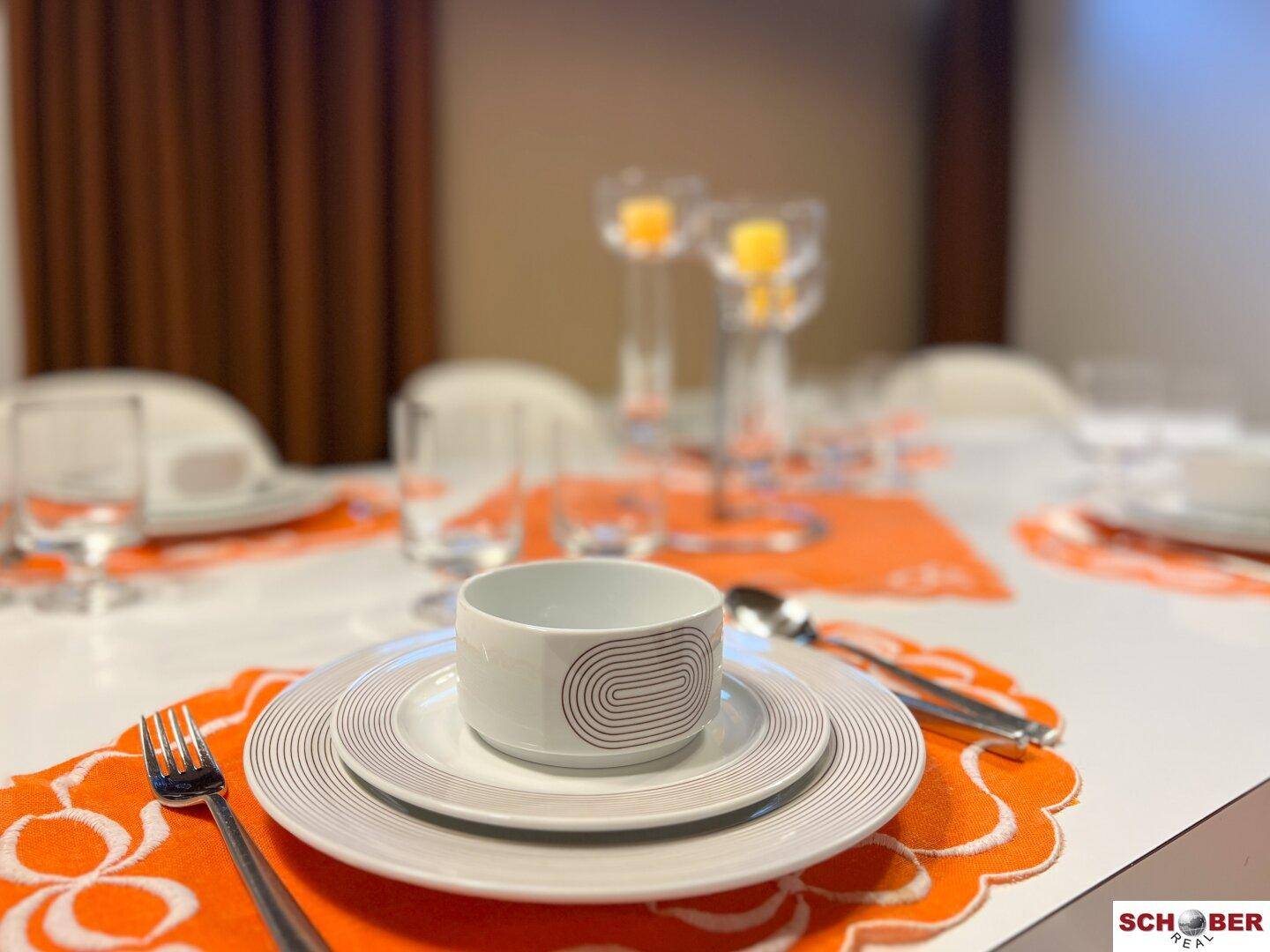 Nahaufnahme eines gedeckten Esstisches mit Tellern, Besteck und orangefarbenen Platzsets.
