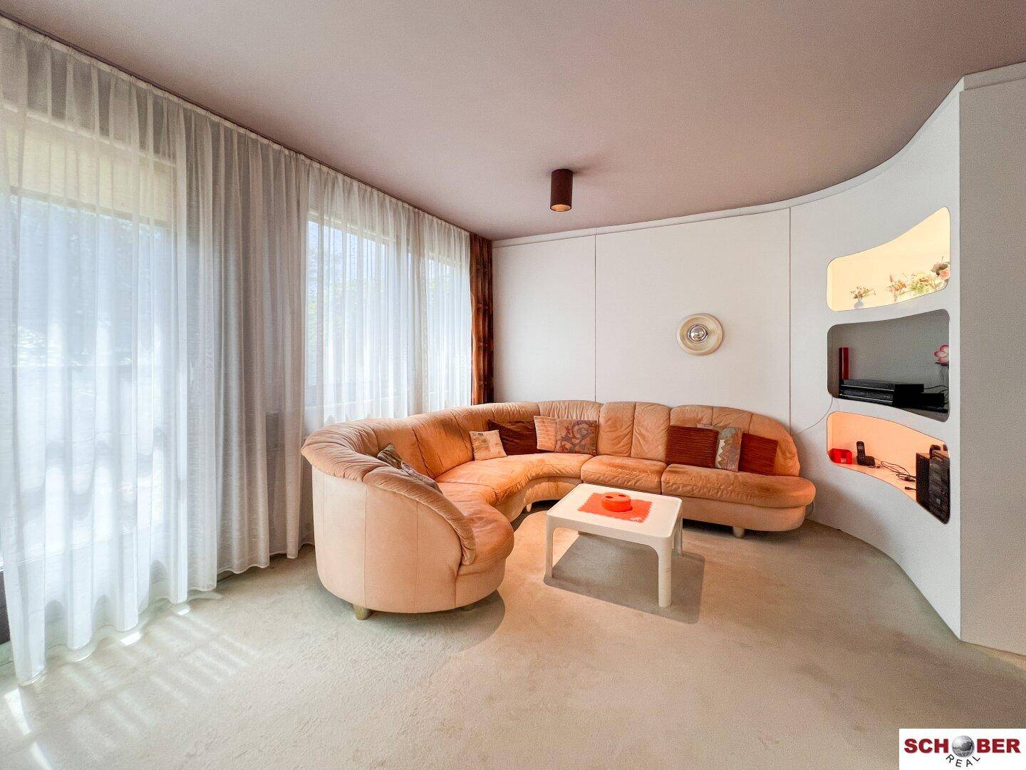 Gemütliches Wohnzimmer mit einem großen, geschwungenen Sofa und hellen Fenstern, die viel Tageslicht hereinlassen.