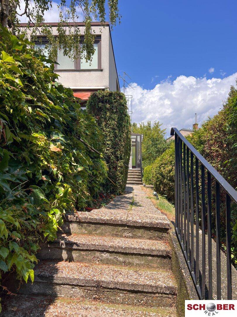 Steintreppe führt nach oben, umgeben von dichten grünen Hecken und einem Gebäude im Hintergrund.