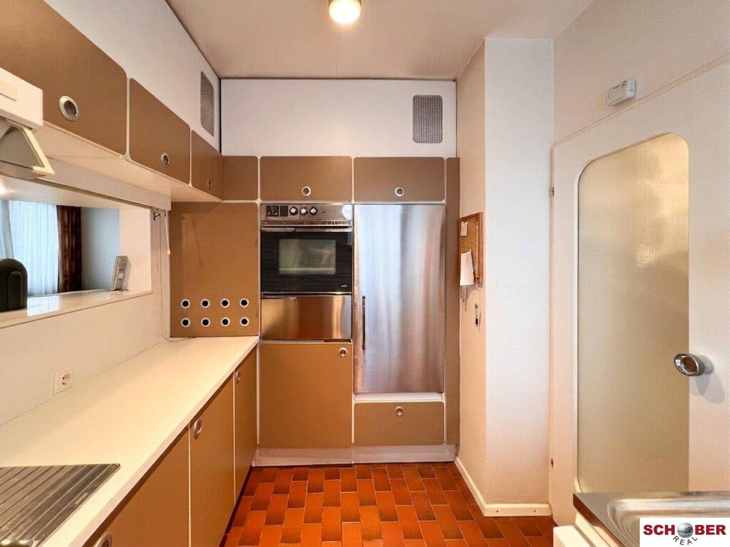 Funktionale Küche mit braunen Einbauschränken, weißen Arbeitsplatten und einem Backofen sowie Kühlschrank.