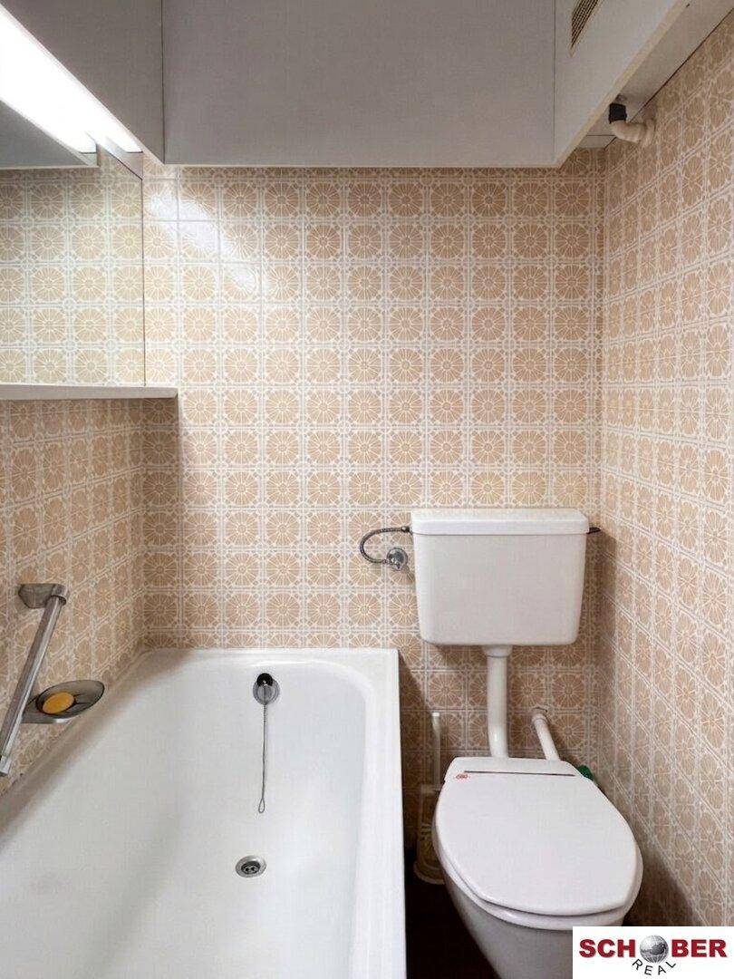 Badezimmer mit Badewanne und Toilette, ausgestattet mit gemusterten beigefarbenen Fliesen.
