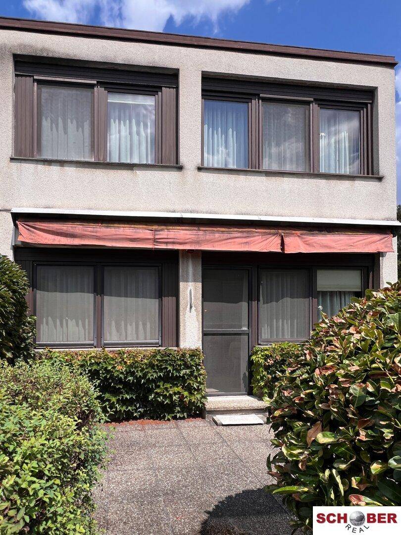 Fassade des Gebäudes mit mehreren Fenstern, einem Eingangsbereich und grünen Sträuchern davor.