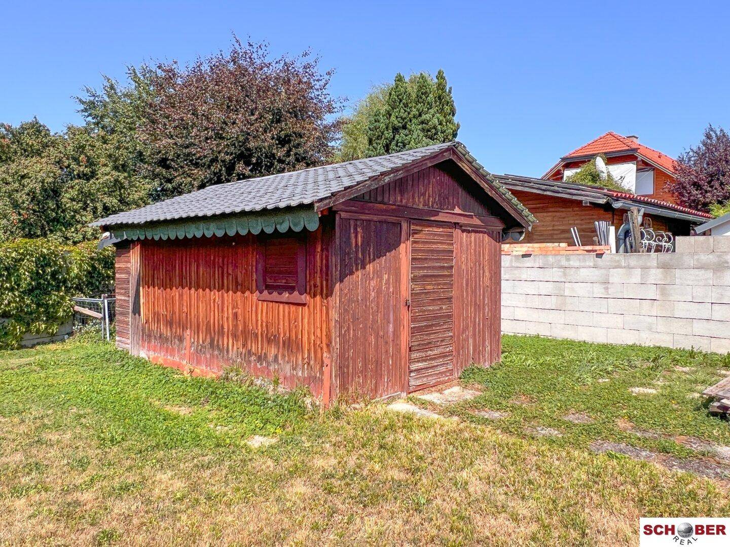 Rustikaler Holzschuppen mit dunklem Dach auf einer Rasenfläche, umgeben von Bäumen und einer Mauer.
