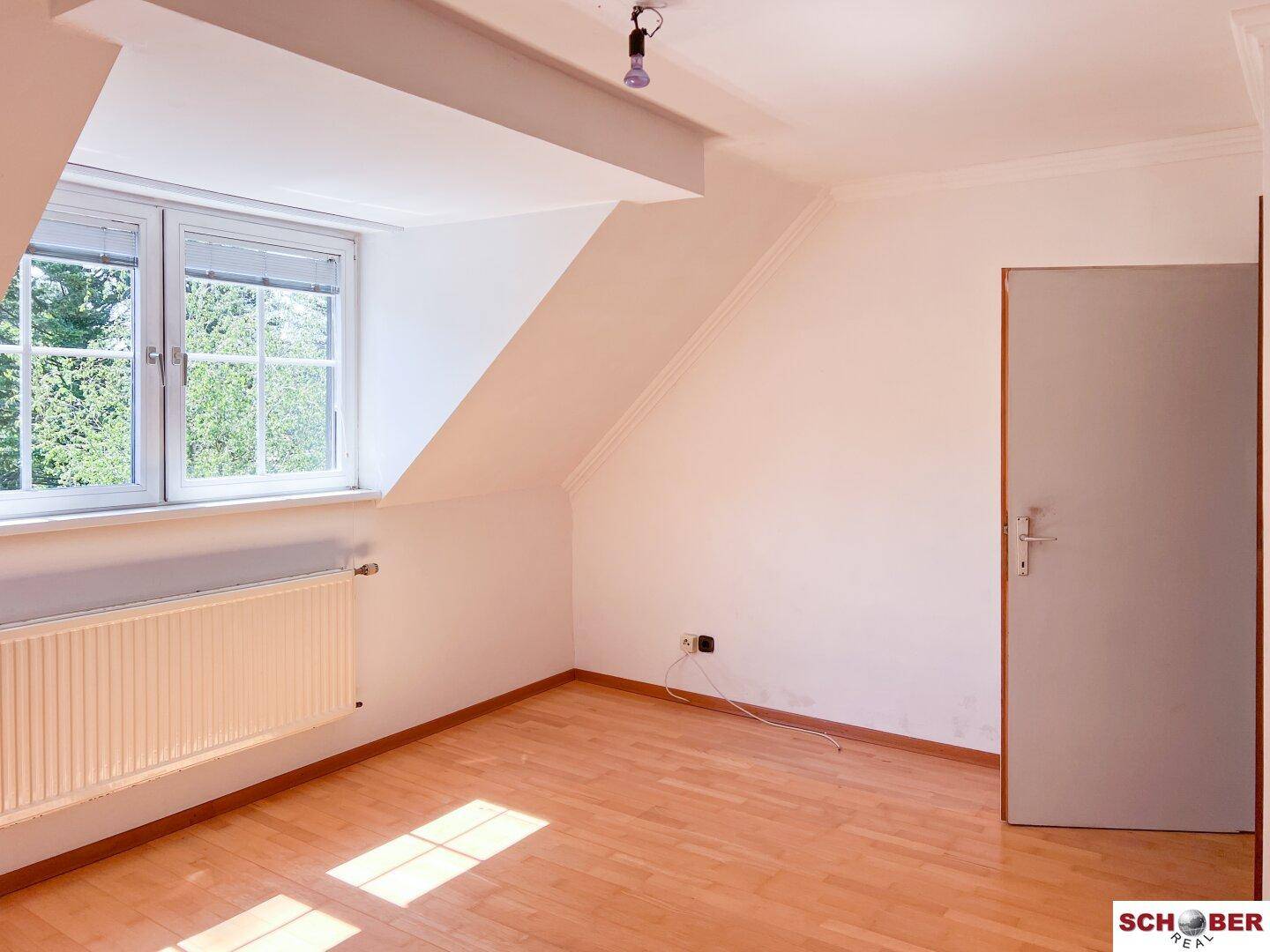 Helles Zimmer im Dachgeschoss mit schrägen Wänden, Holzfußboden und großem Fenster mit Blick ins Grüne.