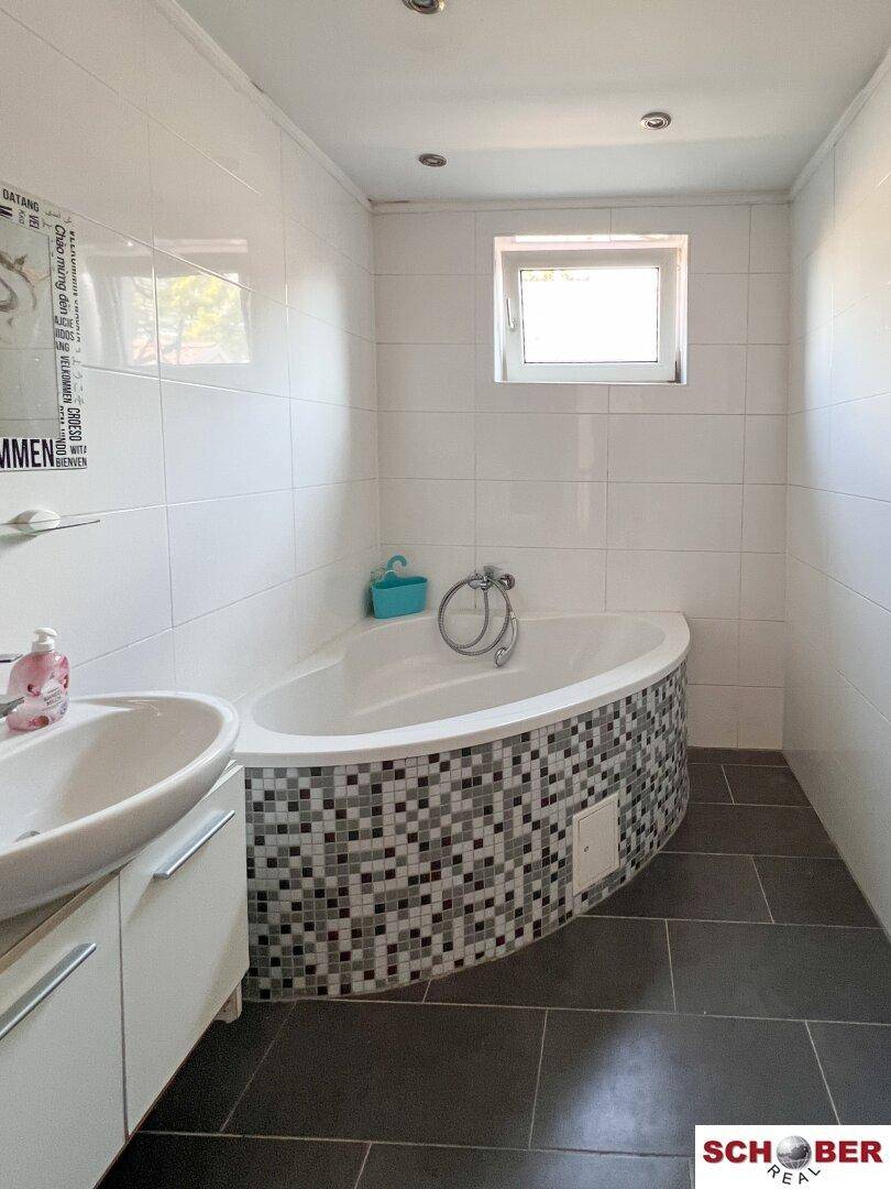 Badezimmer mit Eckbadewanne, weißen Fliesen und einem Fenster für natürliche Belüftung.