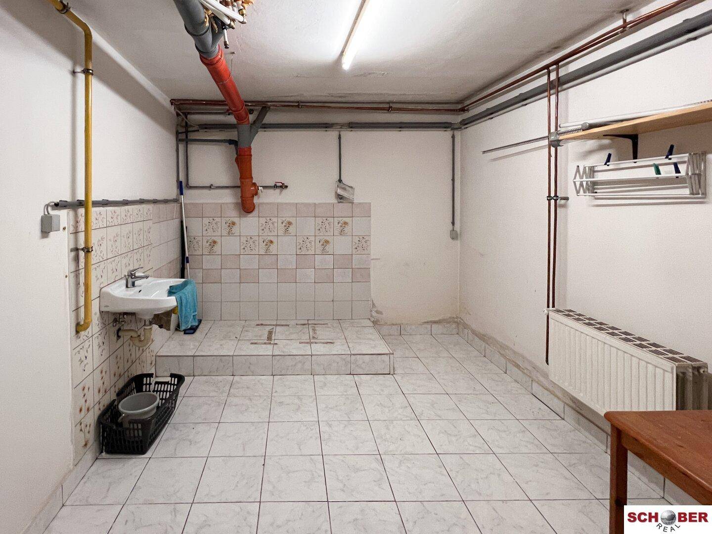 Geräumiger Kellerraum mit Fliesenboden, Waschbecken und sichtbaren Leitungen, ideal als Waschküche.