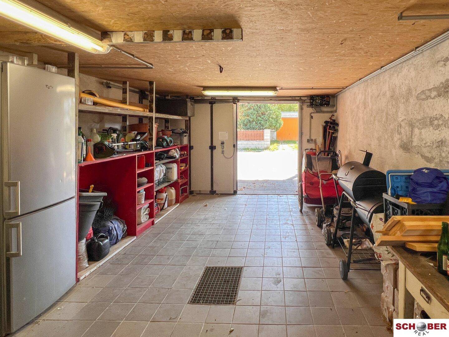 Geräumige Garage mit Fliesenboden, Regalen und einem Kühlschrank, die auch als Abstellraum dient.
