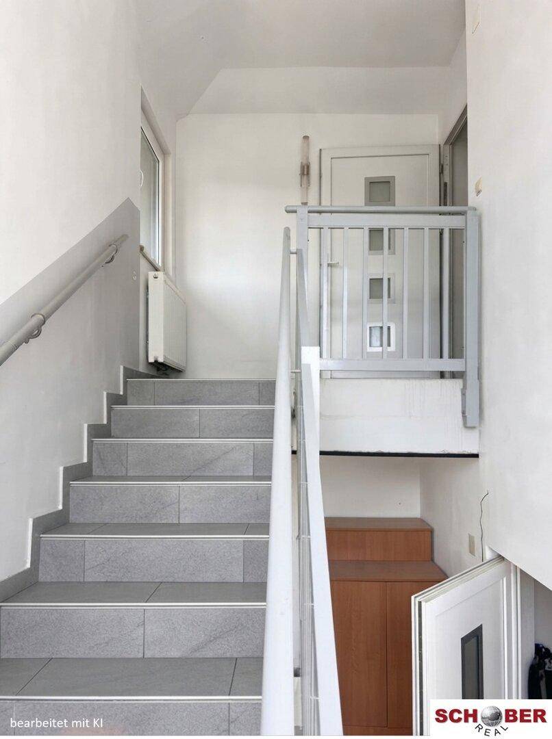 Heller Treppenaufgang mit grauen Fliesen und Handlauf, der in die obere Etage führt.