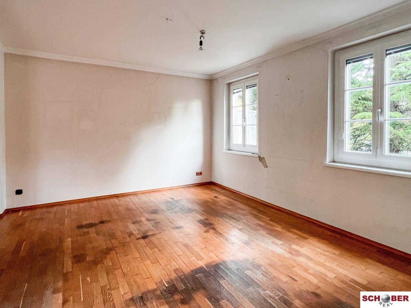 Leeres Schlafzimmer mit Holzboden und zwei Fenstern, die viel Tageslicht hereinlassen.