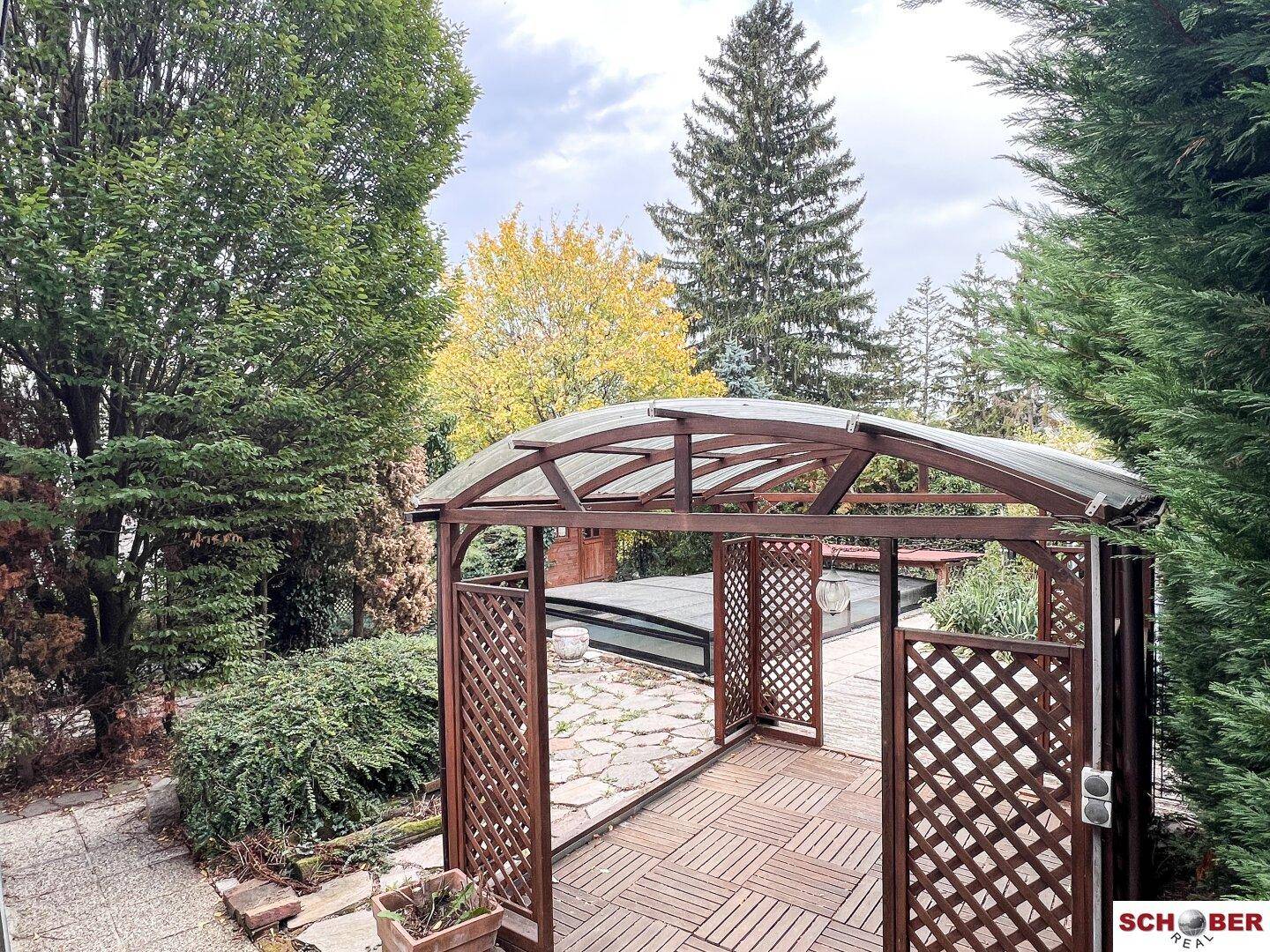Holzpavillon mit Gitterwänden und Blick auf den Garten mit überdachtem Pool und Bäumen.