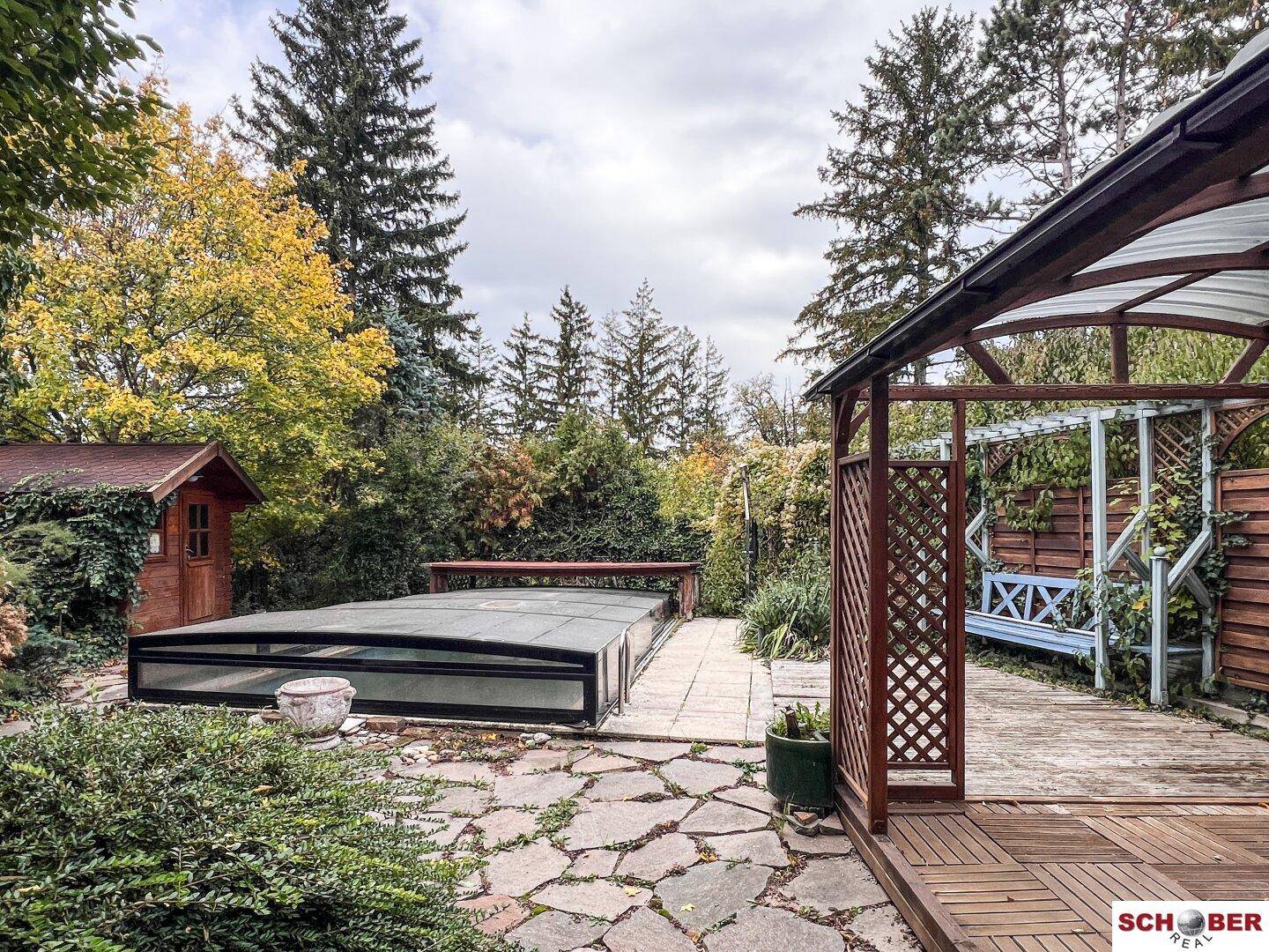 Großer Gartenbereich mit überdachtem Pool, Holzterrasse und einem Pavillon umgeben von Bäumen.