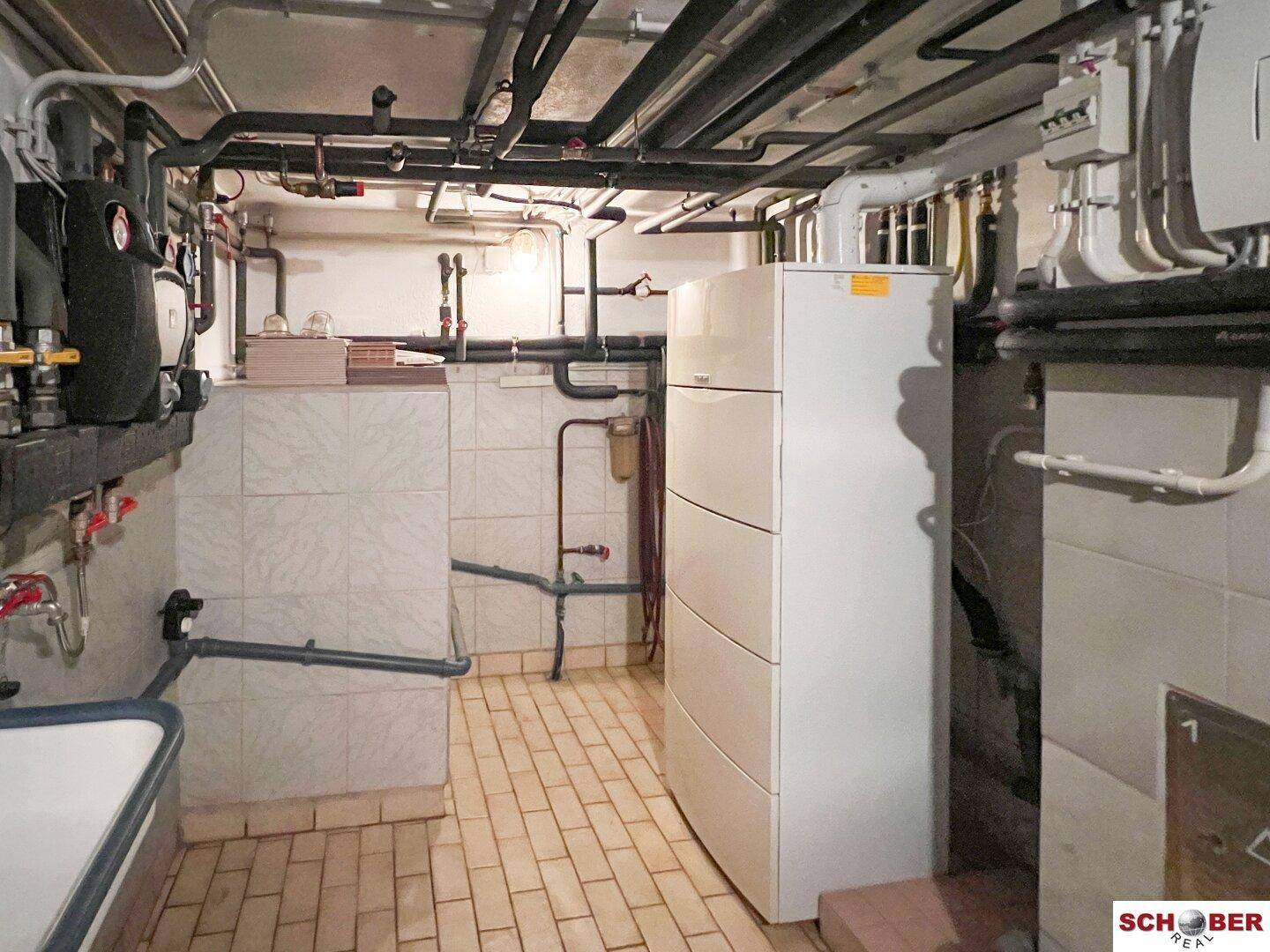 Heizungsraum im Keller mit sichtbaren Rohren, Boiler und Fliesenboden.