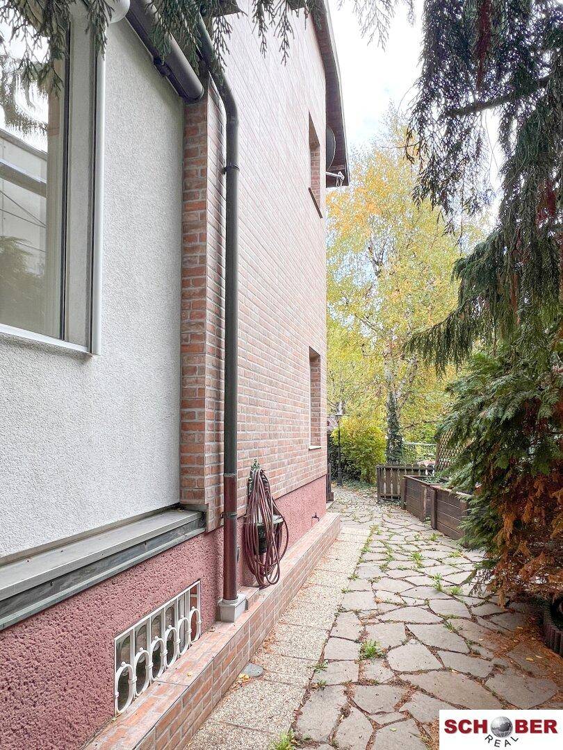 Seitenansicht eines Backsteinhauses mit Fenstern, Fallrohr und einem gepflasterten Weg im Garten.