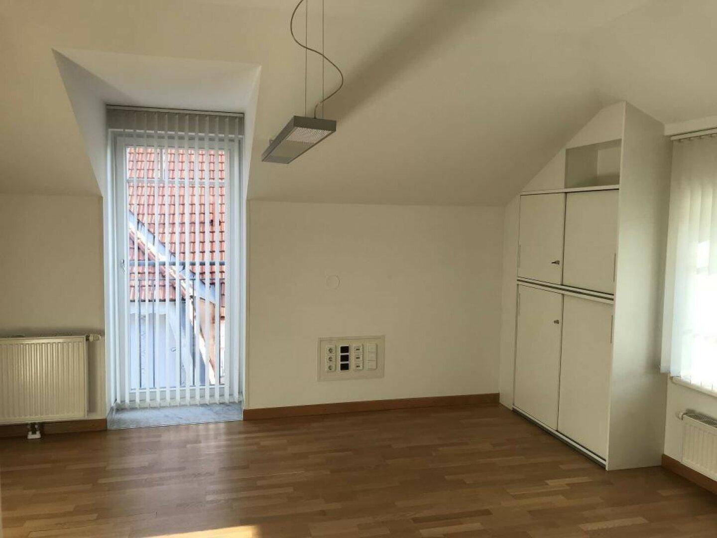 Leerer Raum im Dachgeschoss mit Dachschräge, hohem Fenster und integriertem weißen Schrank.