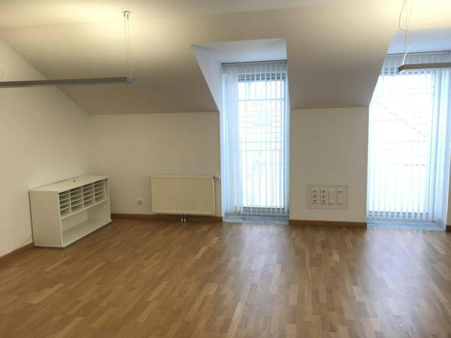 Leerer Raum im Dachgeschoss mit zwei hohen Fenstern, Holzboden und niedrigem Schrank.