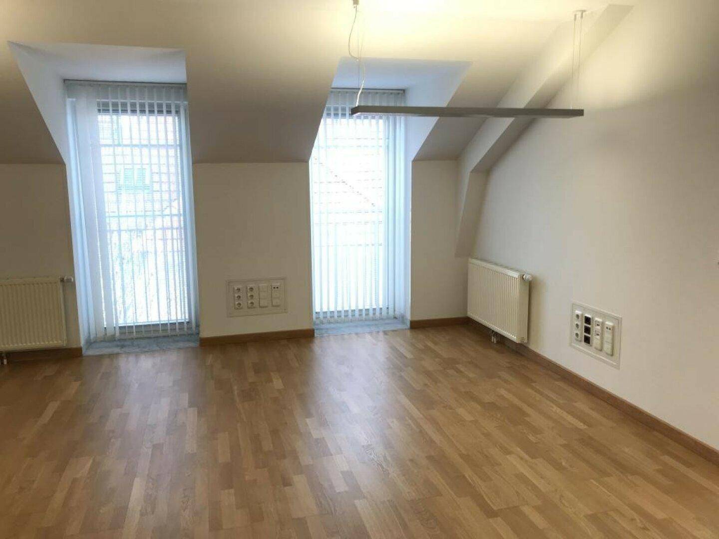 Leerer Raum im Dachgeschoss mit Dachschräge, zwei hohen Fenstern und Holzboden.