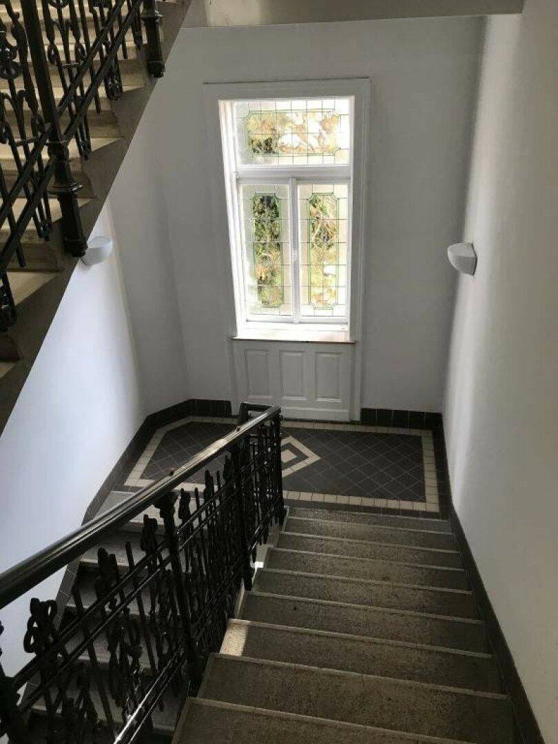Treppenhaus mit schmiedeeisernem Geländer und einem Buntglasfenster, das Tageslicht spendet.