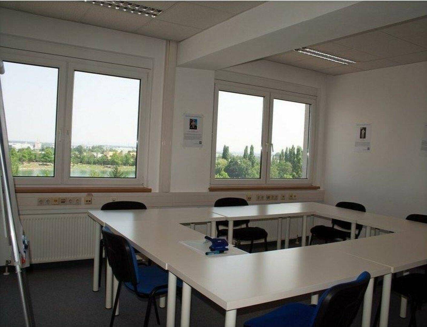 Konferenzraum mit U-förmigem Tisch, Stühlen und großen Fenstern mit weitem Blick ins Grüne.