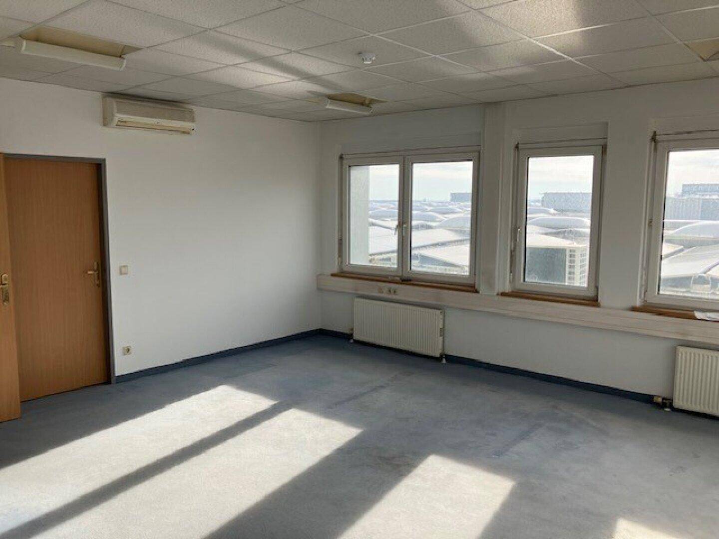Geräumiges Büro mit blauem Teppichboden, Klimaanlage und Blick auf die umliegenden Gebäude.