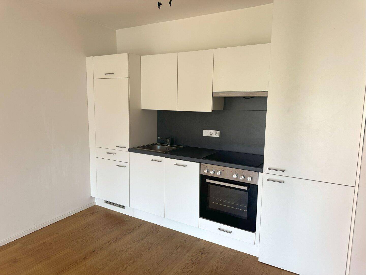 Moderne, kompakte Küchenzeile mit weißen Schränken, integriertem Backofen und Kochfeld auf Parkettboden.